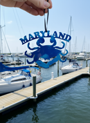 Blue 'Maryland' crab ornament held over a marina with boats in the background.
