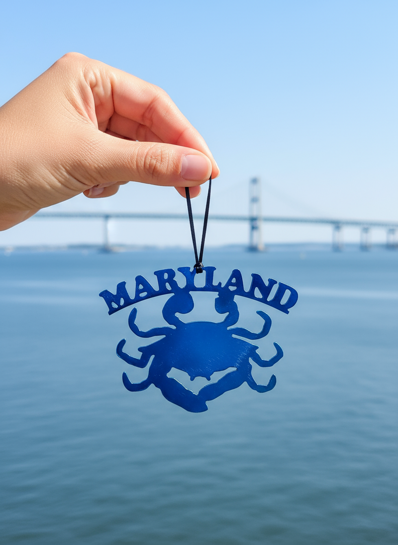 Hand holding a blue 'Maryland' ornament with a crab design against a blue sky and water background.